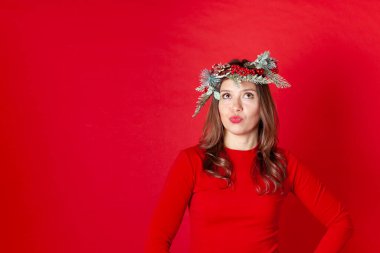 Noel çelengi giymiş düşünceli genç bir kadının portresi, kırmızı bir arka planda, ördek gibi dudaklar yukarı bakıyor.