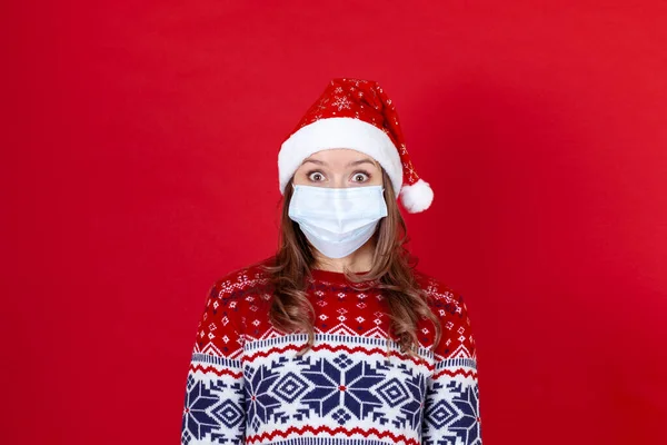 Una joven con una máscara médica, sombrero de Santa Claus rodea sus ojos de horror o sorpresa sobre un fondo rojo —  Fotos de Stock
