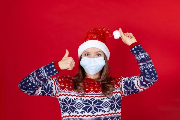 Una mujer joven con una máscara médica, sombrero de Santa Claus levanta el pulgar hacia arriba, aislado sobre un fondo rojo con espacio para el texto —  Fotos de Stock