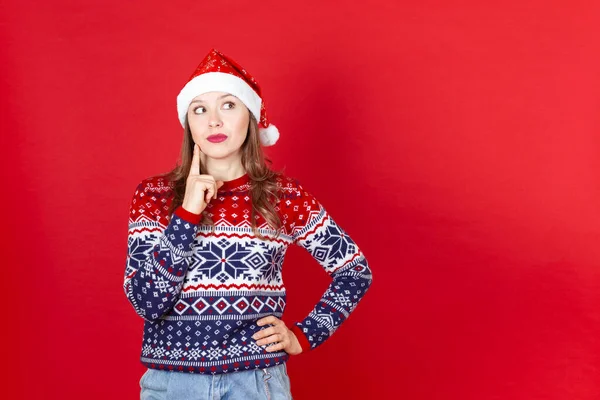 クリスマスセーターのいたずら好きな若い女性とサンタの帽子が横に見える、赤い背景にスペースをコピー — ストック写真