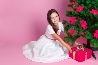 Uzun saçlı, beyaz elbiseli, gülümseyen kız pembe arka plandaki Noel ağacının altından hediye kutusunu aldı.