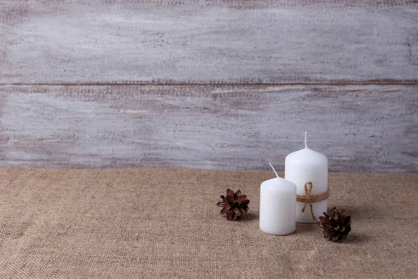 mock up of two white candles in jute rope, cones on a wooden background and burlap
