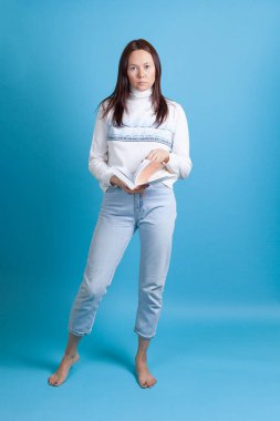 Uzun boylu, ince Asyalı, süveter ve kot pantolonlu genç bir kadın mavi arka planda izole edilmiş bir kitap okuyor.