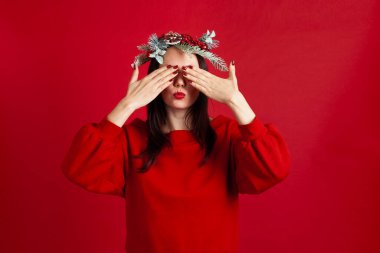 Asyalı genç bir kadının yakın plan portresi. Çelenk takmış, elleriyle gözlerini kapatmış ve kırmızı arka planda öpücük yollamış.