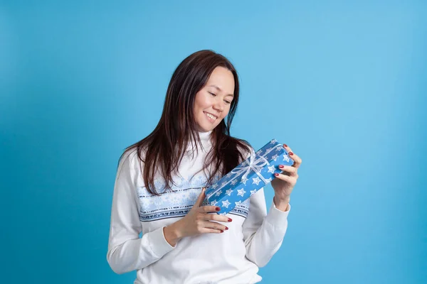 Mock up glimlachen aziatische jonge vrouw in een trui met sneeuwvlokken kijkt naar een geschenk doos geïsoleerd op een blauwe achtergrond — Stockfoto