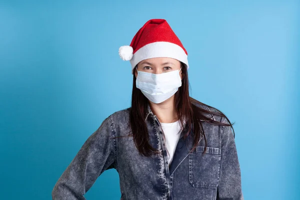 Burla de mujer joven asiática con un sombrero de Santa Claus y una máscara protectora médica de covid-19 sobre un fondo azul —  Fotos de Stock