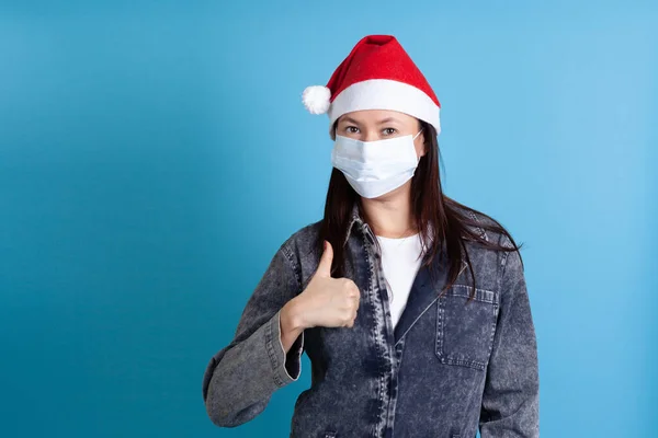 Mujer asiática con un sombrero de Santa Claus y una máscara protectora médica covid-19 mostrando un pulgar hacia arriba sobre un fondo azul —  Fotos de Stock