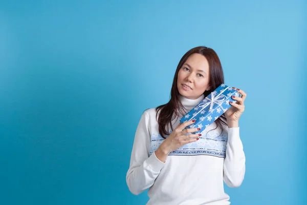 Glimlachende aziatische jong vrouw in een trui met sneeuwvlokken shakes een geschenk doos in de buurt van haar hoofd, geïsoleerd op een blauwe achtergrond — Stockfoto