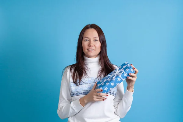 Mock up glimlachen aziatische jonge vrouw in een trui met sneeuwvlokken holding een nieuw jaar cadeau geïsoleerd op een blauwe achtergrond — Stockfoto