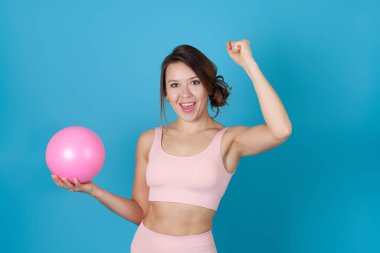 Pembe yoga tulumlu neşeli bir kadının mavi arka planda izole edilmiş bir şekilde elinde top tutan yakın plan portresi.