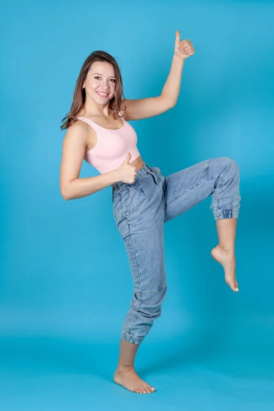 Una mujer risueño de cuerpo entero en jeans y un top rosa baila y da un pulgar hacia arriba aislado sobre un fondo azul — Foto de Stock