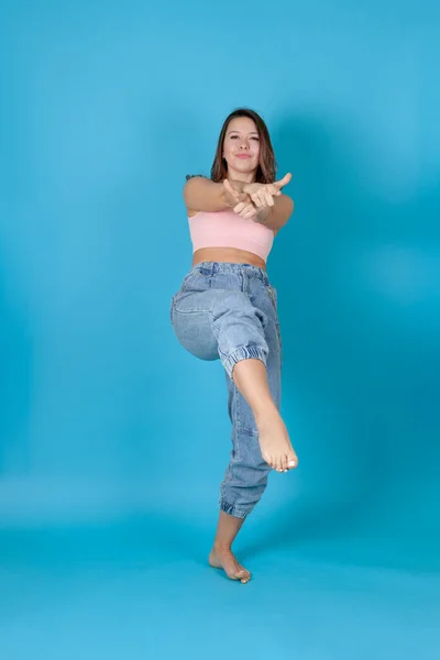 Una mujer feliz de cuerpo entero en jeans y un top rosa baila y cruza sus brazos aislados sobre un fondo azul — Foto de Stock
