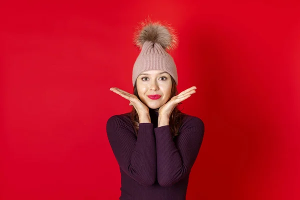 붉은 배경에 고립 된 채, 겨울 모자를 쓰고 손을 그녀의 얼굴에 대고 미소짓고 있는 젊은 여성의 근접 사진 — 스톡 사진