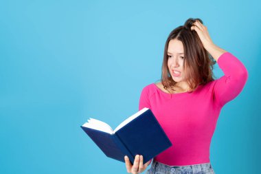 Elinde bir günlük ya da kitap tutan ve hayal kırıklığı içinde saçını çeken genç bir kadının mavi bir arka planda izole edilmiş portresi.