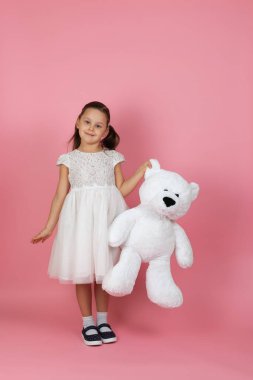 Uzun boylu, at kuyruklu ve beyaz prenses elbiseli yalnız bir kız pembe bir arka planda beyaz bir oyuncak ayıyı kulağından tutuyor.