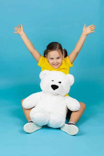 Sonriente, chica encantada con coletas en camiseta amarilla se sienta en el suelo y levanta las manos junto al oso de peluche blanco, aislado sobre un fondo azul — Foto de Stock