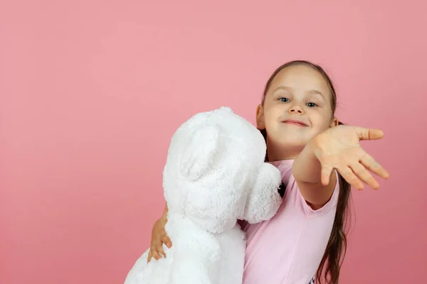 Gros plan une belle fille charmante avec des queues de cheval et une robe rose serre un ours en peluche blanc et nous salue, isolé sur un fond rose — Photo