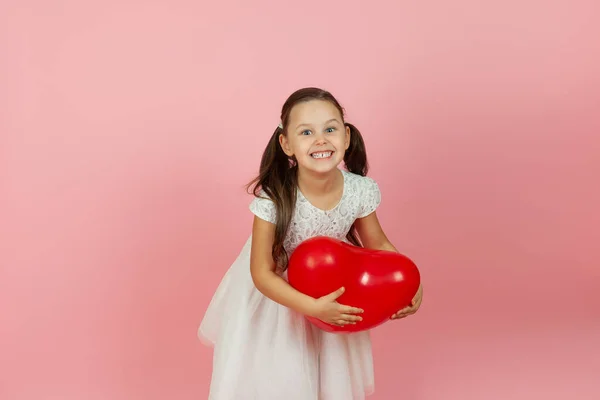 Chica encantada, complacida con colas de caballo y en vestido blanco se para a medias y sostiene el globo rojo en forma de corazón, aislado sobre fondo rosa — Foto de Stock