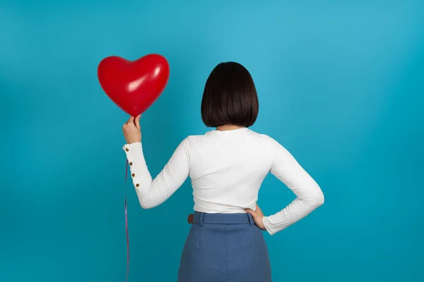 Mujer morena maqueta se para con la espalda y sostiene un globo rojo en forma de corazón, despersonalizado, aislado sobre un fondo azul — Foto de Stock