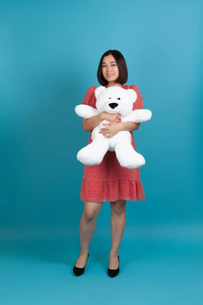 Full-length portret van een glimlachende jonge Aziatische vrouw omarmen een witte teddybeer als een Valentijnsdag geschenk, geïsoleerd op een blauwe achtergrond — Stockfoto