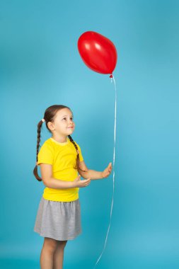 Sarı tişörtlü, at kuyruklu, sevimli bir kız kırmızı bir kalp şeklinde balonu ipin ucunda tutuyor, mavi arka planda izole edilmiş.
