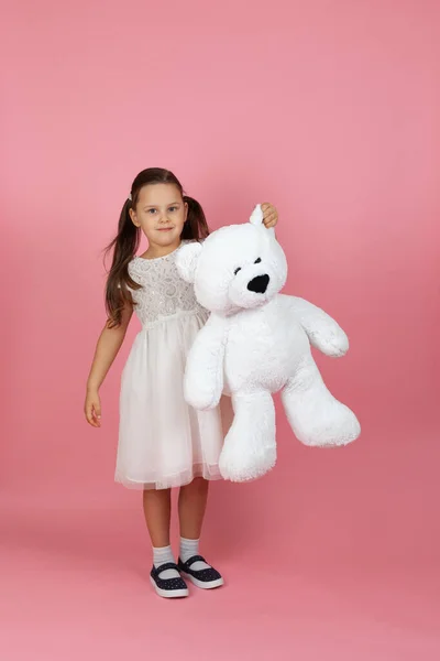 Full-length lachend schattig meisje met paardenstaarten en in een witte prinses jurk houdt een witte teddybeer door het oor, geïsoleerd op een roze achtergrond — Stockfoto