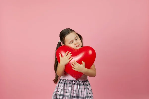 Primer plano chica suave en vestido rosa con los ojos cerrados abraza un globo rojo en forma de corazón a la cabeza, aislado sobre un fondo rosa — Foto de Stock