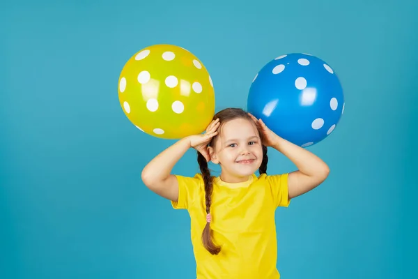 Niña riendo de cerca con coletas en una camiseta amarilla sostiene globos en su cabeza como orejas, aislados sobre un fondo azul —  Fotos de Stock