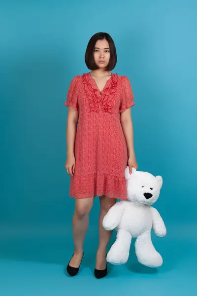 Full-length portret van een verdrietig, boos, beledigd, eenzame vrouw in een rode jurk met een witte teddybeer aan het oor, geïsoleerd op een blauwe achtergrond — Stockfoto