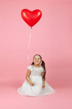 At kuyruklu ve beyaz elbiseli mutlu çocuk yerde oturuyor, üzerinde kırmızı kalp şeklinde bir balon uçuşuyor, pembe bir arka planda izole edilmiş.