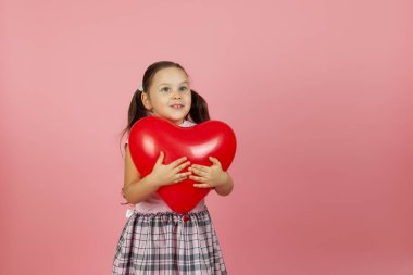 Şaşırmış, mutlu pembe elbiseli bir kız kırmızı bir kalp şeklindeki balona sarılıyor ve pembe bir arka planda soyutlanmış bir şekilde yana bakıyor.