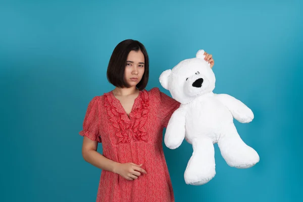 Close-up portret van een verdrietig, boos, beledigd, teleurgesteld vrouw in een rode jurk met een witte teddybeer aan het oor, geïsoleerd op een blauwe achtergrond — Stockfoto