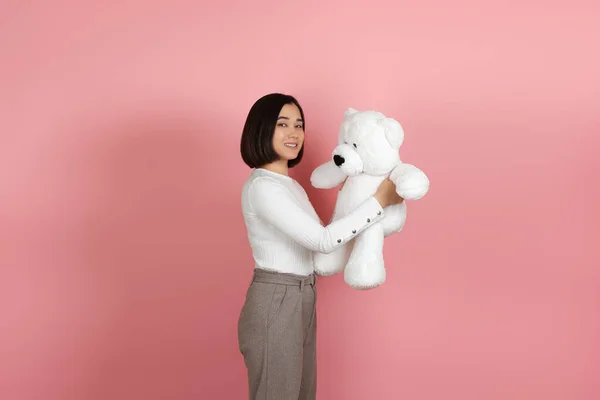 Studio portret van een glimlachende mooie Aziatische vrouw staan zijwaarts en het houden van een witte teddybeer geïsoleerd op een roze achtergrond — Stockfoto