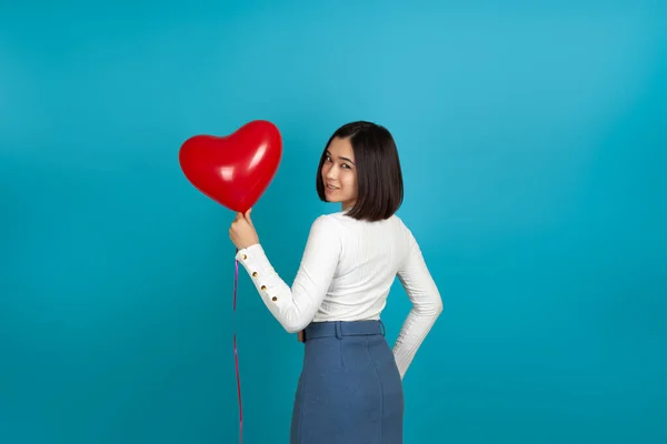 Aziatische vrouw draait haar rug naar de camera en houdt een rode hartvormige ballon geïsoleerd op een blauwe achtergrond — Stockfoto