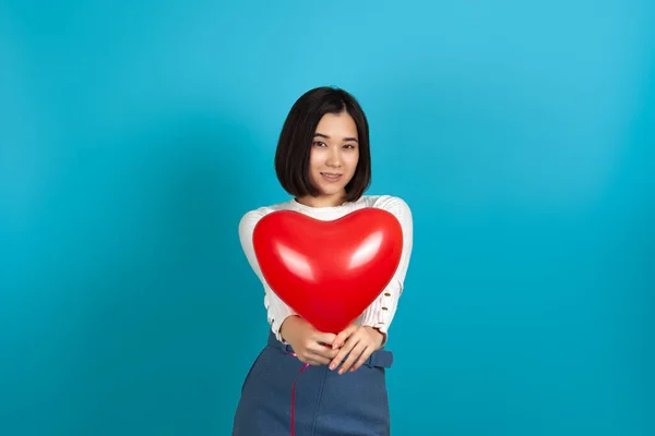 Mock up een glimlachende aziatische vrouw houdt een rode hartvormige ballon geïsoleerd op een blauwe achtergrond naar de camera — Stockfoto