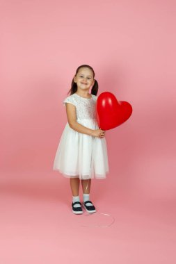 Tam boy dikkatsiz, ilgisiz, beyaz elbiseli kız pembe arka planda izole edilmiş kırmızı kalp şeklinde balon tutuyor.