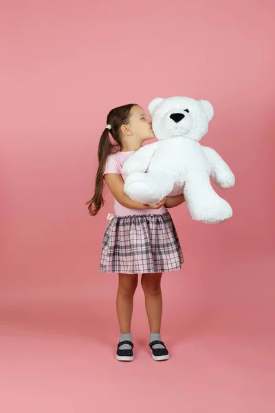 Full-length grappig meisje met paardenstaarten en een roze jurk houdt een witte teddybeer en kust het op de wang, geïsoleerd op een roze achtergrond — Stockfoto