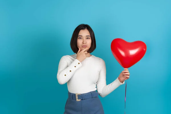 Pensive frons serieus denken aziatische vrouw met de hand onder kin, houden rode hartvormige ballon geïsoleerd op blauwe achtergrond — Stockfoto