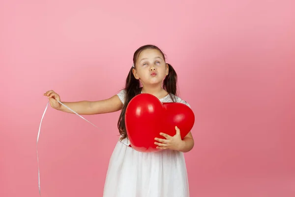 Primer plano chica en un vestido blanco rueda los ojos y sostiene un globo rojo en la forma de un corazón y envía un beso aislado sobre un fondo rosa — Foto de Stock