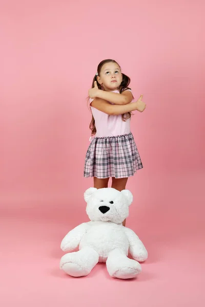 Volledige lengte duur, dromerig meisje met paardenstaarten en roze jurk geeft een duim omhoog met een witte teddybeer in de buurt van haar voeten, geïsoleerd op een roze achtergrond — Stockfoto