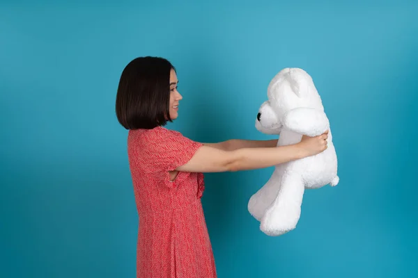 Profilde kırmızı elbiseli gülümseyen Asyalı bir kadın kol mesafesinde tuttuğu beyaz bir oyuncak ayıya bakıyor. — Stok fotoğraf