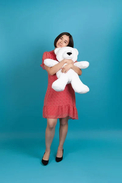 Full-length portret van een glimlachende jonge Aziatische vrouw zachtjes knuffelen een witte teddybeer, geïsoleerd op een blauwe achtergrond — Stockfoto