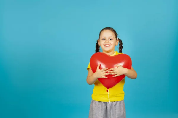 Primer plano alegre, chica entusiasta con coletas en una camiseta amarilla abraza un globo rojo en forma de corazón, aislado sobre un fondo azul — Foto de Stock