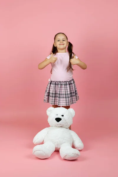 Full-length tevreden, zorgeloos meisje met paardenstaarten en roze jurk geeft duimen omhoog met witte teddybeer in de buurt van haar voeten, geïsoleerd op een roze achtergrond — Stockfoto