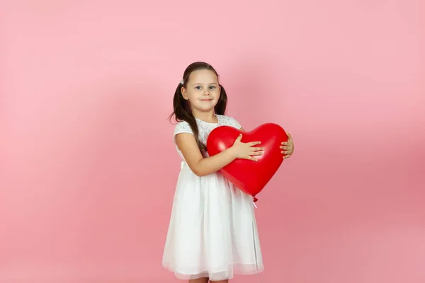 Dulce niña en un vestido blanco se para a medias y sostiene un globo rojo en forma de corazón en el día de San Valentín, aislado sobre un fondo rosa — Foto de Stock