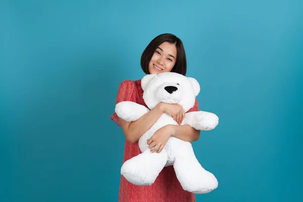 Close-up portret van een glimlachende jonge Aziatische vrouw in een rode jurk knuffelen een witte teddybeer in de buurt van haar borst, geïsoleerd op een blauwe achtergrond — Stockfoto