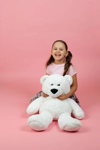 Een vrolijk, lachend meisje met paardenstaarten en een roze jurk zit op de vloer naast een witte teddybeer, geïsoleerd op een roze achtergrond — Stockfoto