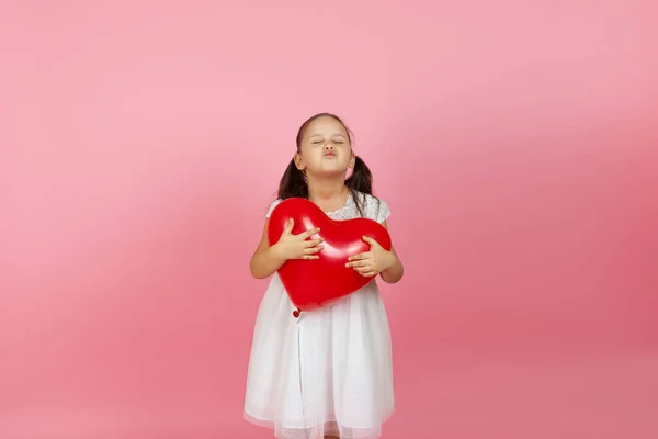 Una chica con los ojos cerrados en un vestido blanco sostiene un globo rojo en forma de corazón y sopla un beso, aislado sobre un fondo rosa — Foto de Stock