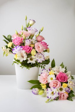 Farklı boyutlarda iki buket gül, papatya, lisianthus, kasımpatı, açılmamış tomurcuklar beyaz bir arka planda beyaz bir karton kutuda, bir buket masanın üstünde.
