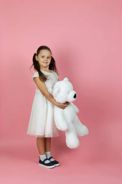 Een volledig lengte ondeugend, mooi meisje met paardenstaarten en een witte prinses jurk houdt een witte teddybeer geïsoleerd op een roze achtergrond — Stockfoto